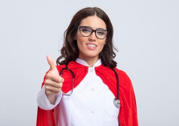 Supermulher sorridente em uniforme de médico com capa vermelha e estetoscópio em óculos ópticos polegares para cima isolado na parede branca