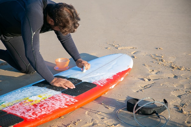 Surfista masculino com roupa de neoprene polindo prancha de surfe com cera na areia