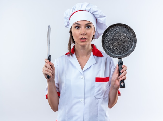 Surpreendeu a jovem cozinheira vestindo uniforme de chef segurando uma frigideira com cutelo isolado na parede branca