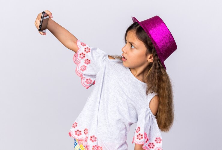 Foto grátis surpreendeu a menina caucasiana com chapéu de festa roxo olhando para o telefone tirando selfie isolada na parede branca com espaço de cópia