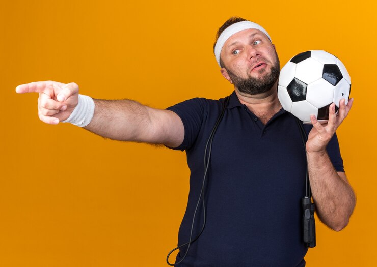 Foto grátis surpreendeu o homem adulto eslavo desportivo com pular corda em volta do pescoço usando bandana e pulseiras segurando uma bola e apontando para o lado isolado na parede laranja com espaço de cópia