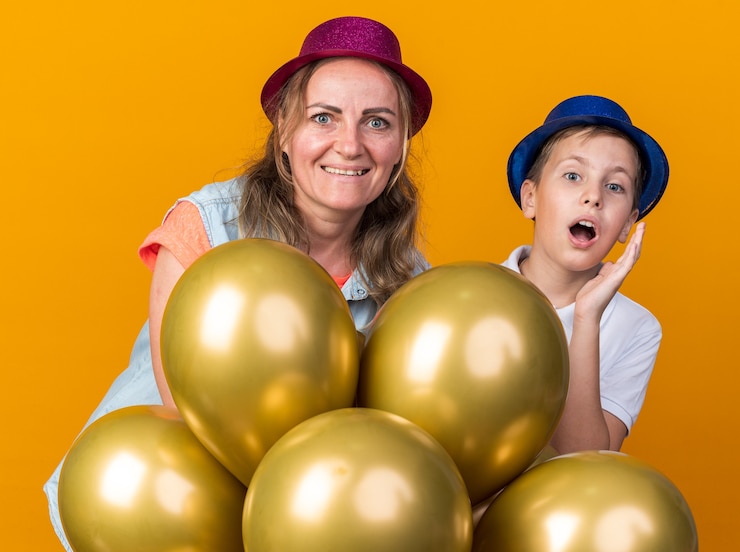 Foto grátis surpreendeu o jovem eslavo com um chapéu de festa azul segurando balões de hélio com sua mãe usando um chapéu de festa roxo isolado na parede laranja com espaço de cópia