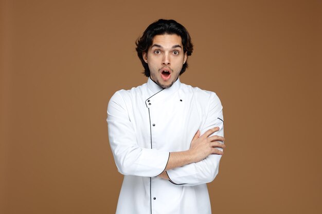 surpreendido jovem chef masculino vestindo uniforme mantendo os braços cruzados olhando para câmera isolada em fundo marrom