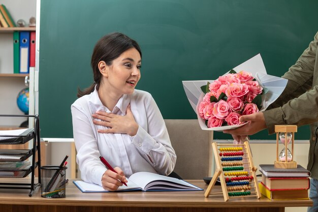 Surpresa, colocando a mão no coração, jovem professora escreve no caderno dá buquê por alguém sentado à mesa com ferramentas escolares na sala de aula