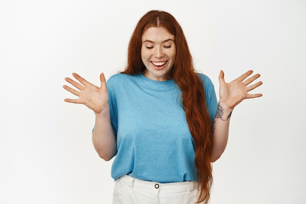 Surpresa. garota ruiva feliz comemorando, apertando as mãos e olhando para baixo com expressão alegre e animada, ganhando algo, presente aberto, de pé contra um fundo branco.