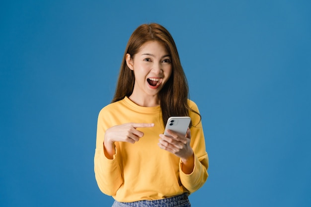Surpresa jovem asiática usando telefone celular com expressão positiva, amplamente sorri, vestida com roupas casuais e olhando para a câmera sobre fundo azul. Mulher feliz adorável feliz alegra sucesso.