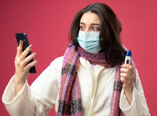 Foto grátis surpresa jovem caucasiana doente vestindo manto e lenço com máscara segurando o telefone celular e termômetro olhando para o telefone isolado na parede carmesim