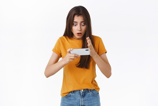 Surpresa jovem chocada e aliviada segurando o smartphone horizontalmente assistindo seu programa de TV favorito via wifi olhar fixo na tela do celular ofegante queixo caído sacudiu ver cliffhanger inesperado