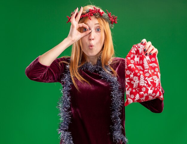 surpresa jovem linda com um vestido vermelho com grinalda e guirlanda no pescoço segurando uma sacola de Natal, mostrando gesto de olhar isolado na parede verde