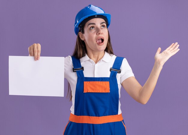 Surpresa olhando para uma jovem construtora de uniforme segurando papel espalhando a mão isolada na parede roxa