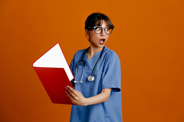 Surpresa segurando pasta jovem médica usando estetoscópio uniforme de quinto isolado em fundo laranja