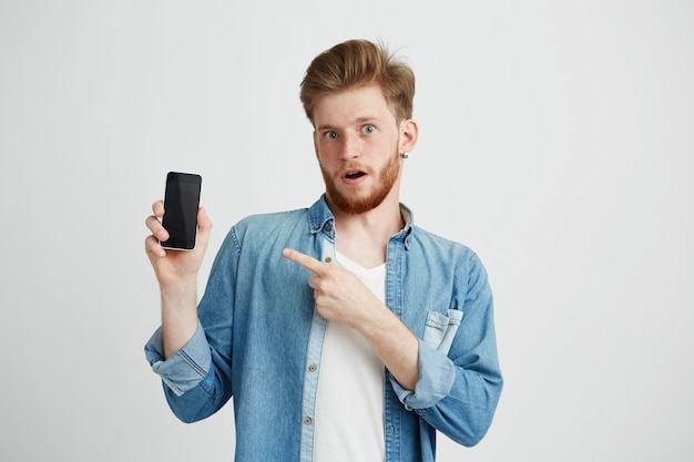 Surpreso jovem bonito com a boca aberta, apontando o dedo para o telefone na mão.