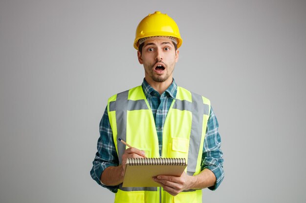 Surpreso jovem engenheiro masculino usando capacete de segurança e uniforme segurando o bloco de notas e lápis olhando para câmera isolada no fundo branco