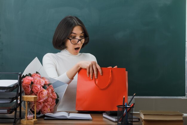 surpreso segurando e olhando para presente jovem professora sentada na mesa com ferramentas escolares em sala de aula