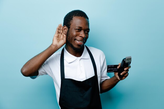 Suspeita mostrando gesto de escuta segurando cartão com máquina de cortar cabelo jovem barbeiro americano africano de uniforme isolado em fundo azul