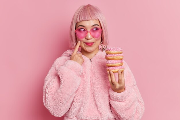 Sweettooth muito sorridente mulher asiática com cabelo rosa na moda usa óculos escuros e casaco de pele detém pilha de donuts posa sonhos interiores sobre algo. Foto monocromática. Mulher gosta de comer donuts