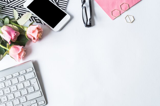 Teclado, smartphone, óculos e rosas cor de rosa em uma superfície branca