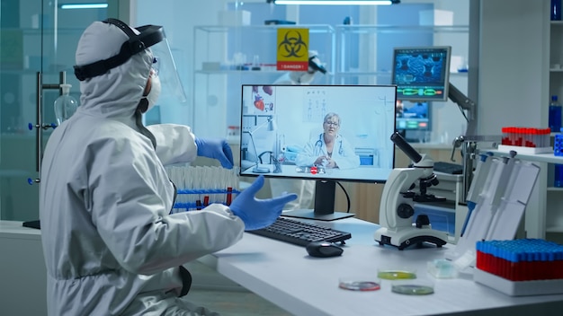 Técnico de laboratório em terno de ppe sentado à mesa no laboratório de pesquisa médica, segurando um tubo de ensaio e conversando em uma videochamada com o médico sênior, dando conselhos on-line durante a reunião virtual