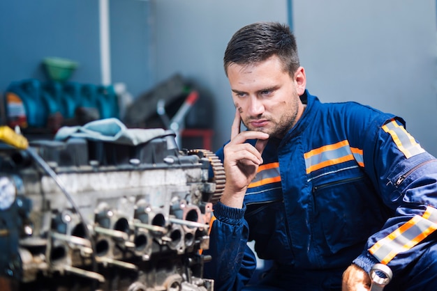 Técnico experiente em mecânico de automóveis, uniformizado, pensando na solução e olhando para o motor do carro na oficina mecânica