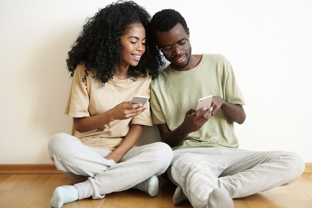 Tecnologia e comunicação modernas. Homem africano atraente com óculos mostrando algo engraçado para a namorada com um corte de cabelo estiloso no celular