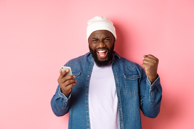 Tecnologia e conceito de compras online. Homem negro feliz regozijando-se, ganhando no app, segurando smartphone e gritando sim, de pé sobre fundo rosa