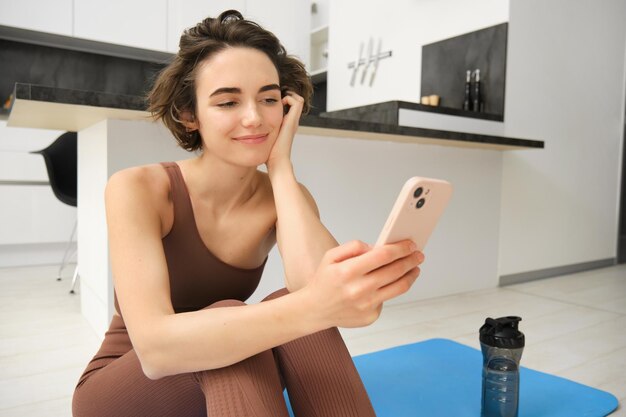 Tecnologia e conceito de esporte garota fitness olhando para seu smartphone sentado em roupas de treino em ho