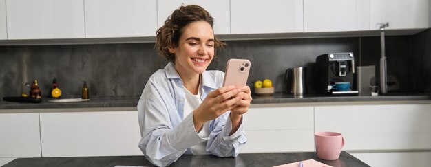Tecnologia e estilo de vida Mulher jovem senta-se em casa usa smartphone em sua cozinha e sorri