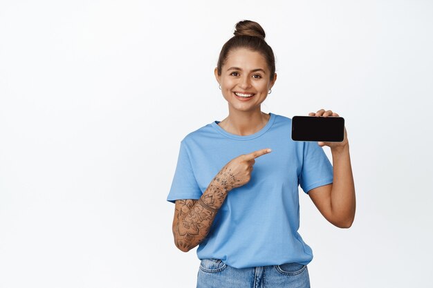 Tecnologia. Mulher sorridente, apontando o dedo na tela horizontal do smartphone, recomendando o aplicativo, loja online, de pé em branco.