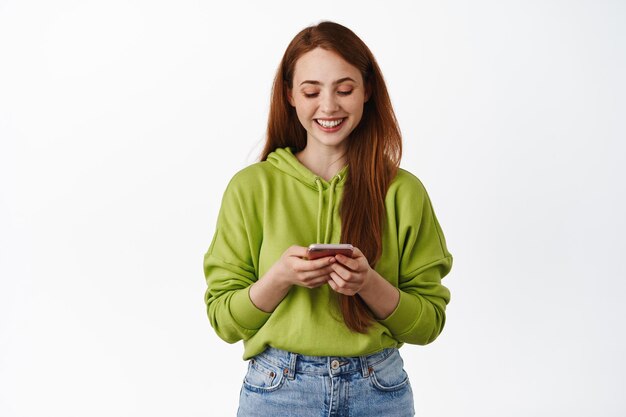 Tecnologia. Ruiva sorridente olhando para smartphone, conversando no telefone, usando o aplicativo no celular, de capuz contra fundo branco.