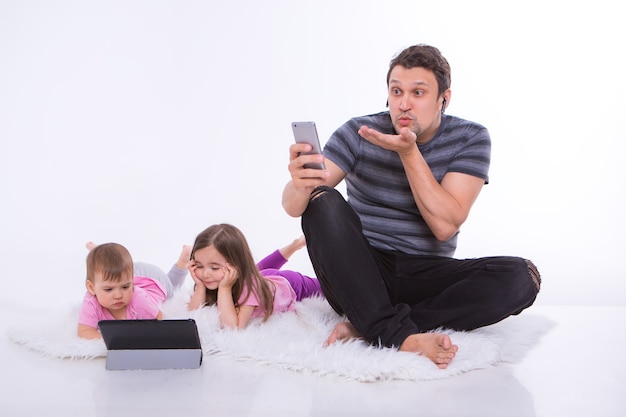 Tecnologias modernas no dia a dia: um homem fala ao telefone por meio de um fone de ouvido, as crianças assistem a um desenho animado em um tablet. hobbies e recreação com gadgets. pai com meninas no chão