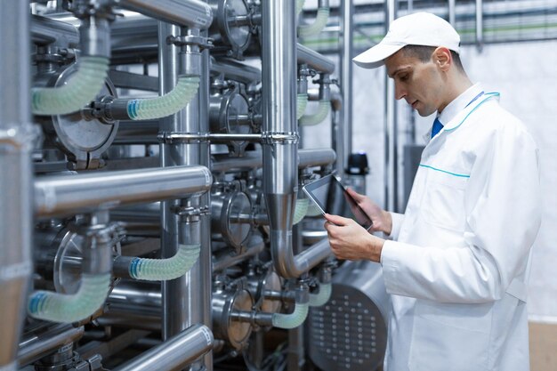 Tecnólogo com tablet cinza nas mãos faz uma configuração da linha de produção em pé no departamento de fábrica de laticínios