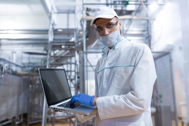 Tecnólogo de jaleco branco com laptop nas mãos controla o processo de produção na loja de laticínios Local para escrever Tecnólogo com laptop está na fábrica