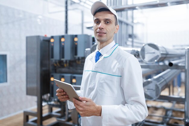 Tecnólogo de jaleco branco faz as entradas necessárias no tablet está na fábrica Um homem com um tablet digital de uniforme no interior da loja de produção do departamento de produção em segundo plano