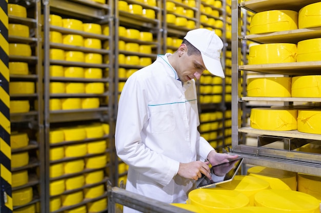 Tecnólogo em uma túnica branca segurando um tablet para registros e fica perto das prateleiras com queijos Produção de produtos de queijo Homem na loja de queijos