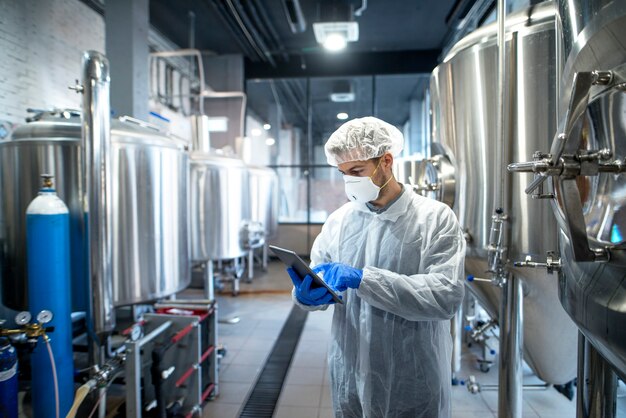 Tecnólogo em uniforme protetor branco controlando o processo industrial usando computador tablet