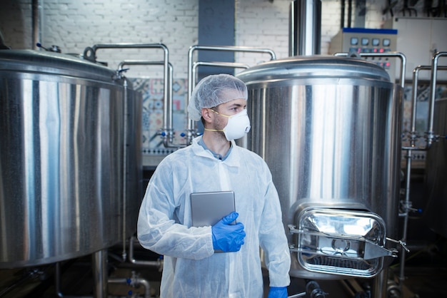 Tecnólogo profissional experiente em uniforme protetor branco segurando o tablet e olhando para o lado na fábrica de produção de alimentos