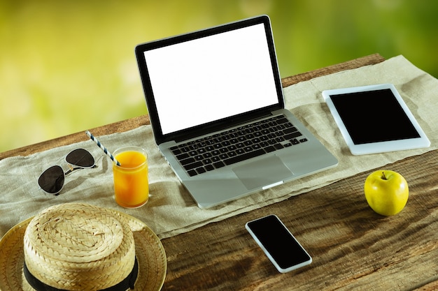 Telas em branco de laptop e smartphone em uma mesa de madeira ao ar livre com a natureza na parede, simulação acima.