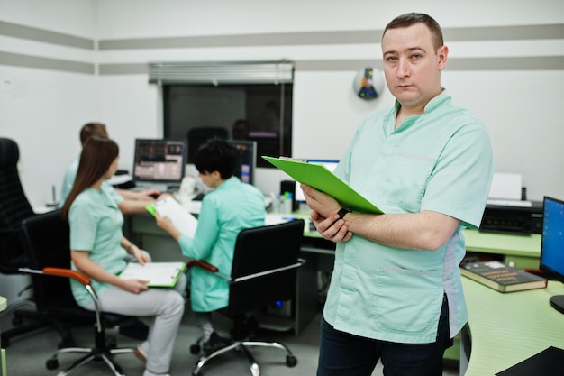 Tema médico retrato de médico masculino com prancheta contra grupo de médicos reunidos no escritório de ressonância magnética no centro de diagnóstico no hospital