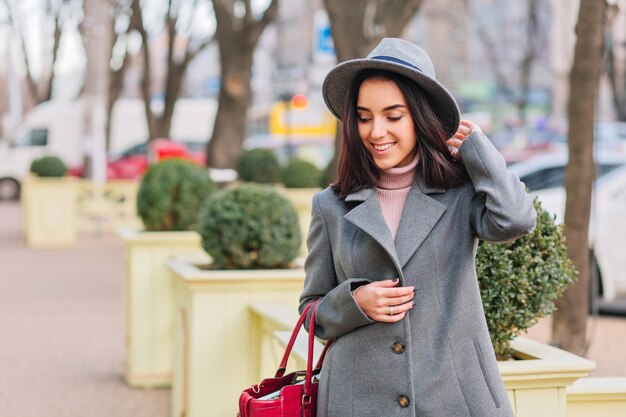 Tempo de caminhada de cidade de alegre elegante jovem de casaco cinza, chapéu, andando na rua na cidade. Sorrindo, expressando verdadeiras emoções faciais positivas, estilo de vida luxuoso e aparência elegante.