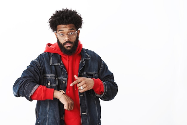 Foto grátis tempo é dinheiro pressa homem barbudo afro-americano masculino intenso confiante com penteado afro em óculos apontando para o relógio como insinuação para se mover mais rápido atrasado para a reunião por cima da parede cinza