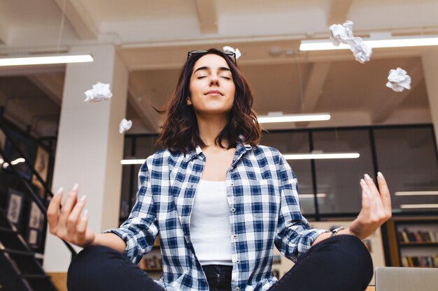 Tempo relaxante alegre jovem morena tendo meditação na mesa no escritório cercam papéis voando. Fazendo uma pausa, pausa, aluno inteligente, relaxamento, grande sucesso, sonhando.