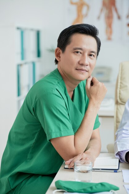 Terapeuta asiática vestindo uniforme verde, sentado na mesa, olhando para o retrato da câmera