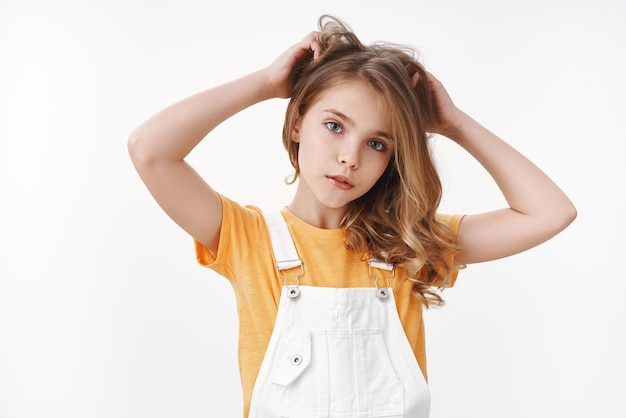 Foto grátis terno e adorável jovem loira com macacão de verão sobre uma camiseta amarela, olhar pensativo e fofo na câmera, arranhar o cabelo incomodado, tocar o penteado, sofrer de piolhos