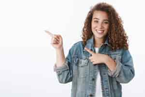 Foto grátis terno feminino jovem aluna penteado encaracolado apontando o dedo indicador esquerdo sorrindo amigavelmente perguntando em que lugar dormitório está alegre parede branca