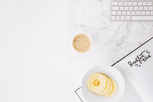 Texto de tempo de pequeno-almoço em papel com teclado; fatias de abacaxi e xícara de café na mesa