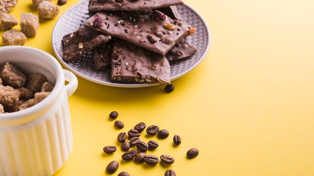 Tigela de açúcar mascavo; grãos de café torrados e barras de chocolate no pano de fundo amarelo