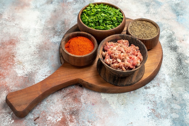 Tigela de vista frontal com tigelas de carne com diferentes especiarias verdes na tábua de cortar na foto de comida de fundo cinza