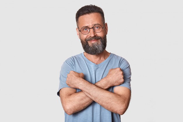 Tipo agradável homem bonito com olhar sincero fica na frente da câmera, tendo um olhar confiante, segurando os braços perto do peito, fazendo cruz, parece satisfeito e engraçado. Conceito de pessoas e emoções.