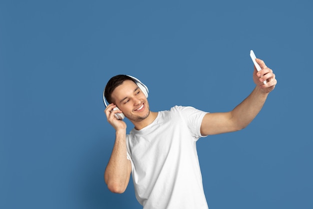 Tirar selfie, ouvir música. Retrato de jovem caucasiano na parede azul do estúdio.