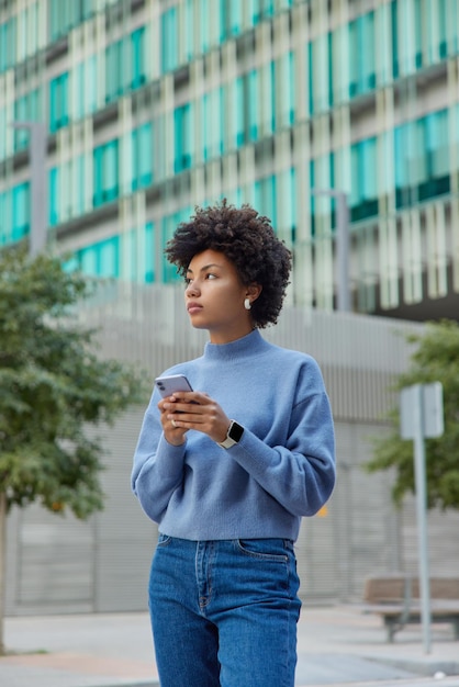 Tiro ao ar livre de mulher bonita pensativa usa jumper azul casual e jeans fica contra o ambiente urbano usa telefone celular conectado à internet sem fio vai chamar passeios de táxi na cidade